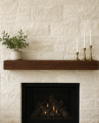 A rustic dark brown wooden mantel shelf mounted above a black fireplace insert, set against a white brick wall flanked by tan brick walls. A small green potted plant and a brass three-arm candlestick holder sit on the mantel.
